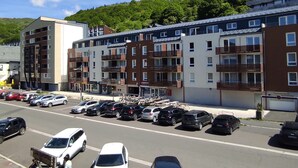 Um edifício residencial com vários andares e varandas, um estacionamento com carros, e uma paisagem acidentada ao fundo.