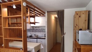 Cozinha compacta com pia, fogão e armários, área de jantar com mesa, e estrutura de madeira com prateleiras.