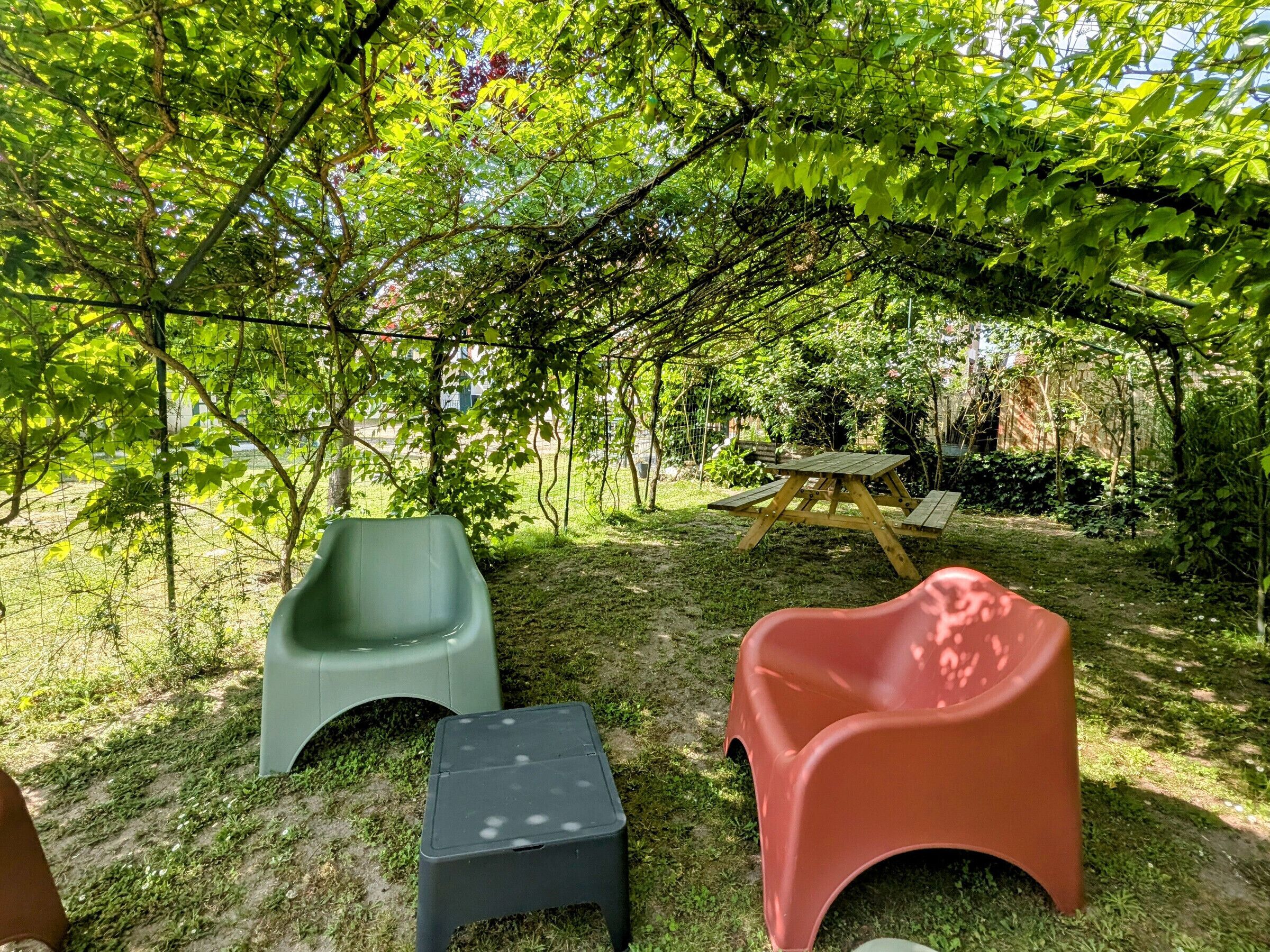 An outdoor seating area with two green and one red plastic chairs, a small black stool, and a wooden picnic table under a tree canopy.