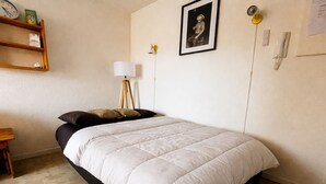 Ein Schlafzimmer mit einem Bett, einem Nachttisch, einer Lampe, einem Regal mit Gegenständen und einem gerahmten Bild an der Wand.