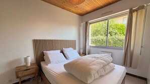 A bedroom with a wooden ceiling, a large window with curtains, a bed with white bedding, and a bedside table with a lamp. - House in Les Sables-d'Olonne with garden, 4 sleeps (Les Sables-d'Olonne)