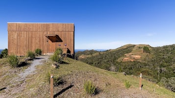 A wooden cabin with a large door and a small window, surrounded by a gravel path and sparse vegetation, overlooking a hilly landscape.