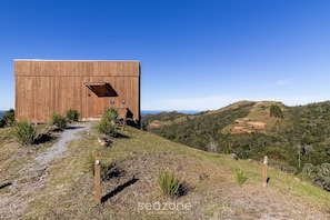 Une cabane en bois, dotée d’une grande porte et d’une petite fenêtre, entourée d’un chemin de gravier et d’une végétation clairsemée, et offrant une vue sur un paysage vallonné.