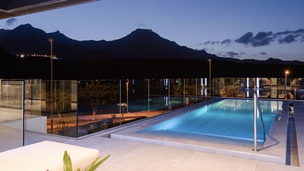A modern outdoor pool area with glass railings, a clear view of mountains in the background, and a well-lit patio area. - Casa Miravella (Adeje)