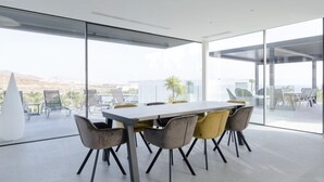 A modern dining area with a large table and chairs, large windows with a view of the landscape, and a glass partition. - Casa Miravella (Adeje)