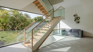 A modern interior with a glass staircase, a grey sofa, and a view of greenery outside. - Casa Miravella (Adeje)