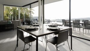 Un comedor moderno con una mesa grande dispuesta para cuatro, ventanas amplias que ofrecen vistas al exterior, y una zona de estar con sofá y sillones.