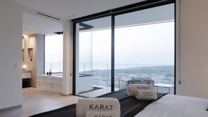 Una habitación de hotel moderna con una cama grande, dos almohadas con la marca 'Karat Luxury Rentals', vistas de un paisaje urbano a través de ventanas del suelo al techo, y un área de baño separada con lavamanos y espejo.