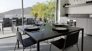 Un comedor moderno con una mesa grande dispuesta para cuatro personas, rodeada de sillas, y vistas a palmeras y montañas a través de amplios ventanales.