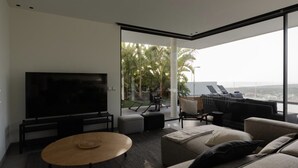 Una sala de estar moderna con un televisor de pantalla plana grande, asientos cómodos y vistas a palmeras y un paisaje urbano a través de ventanas amplias.
