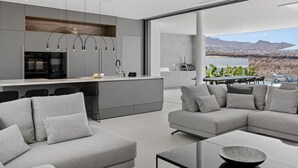 A modern kitchen with a large island, grey sofas, and a dining area with a view of the mountains.