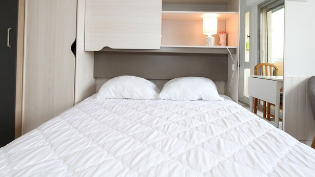 Un lit recouvert d’une courtepointe blanche matelassée, avec deux oreillers, une étagère intégrée au-dessus du lit et un petit bureau avec une chaise, placé  à droite.