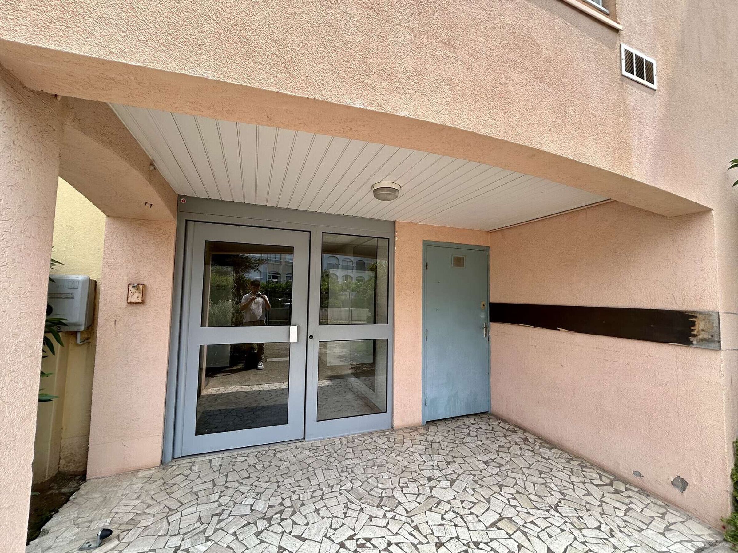 Une entrée d’immeuble avec un patio carrelé, une porte en verre et une porte bleue.