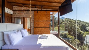 A modern bedroom with a large bed, a brick wall, and a view of a forest. - Cabana romântica vista incrível VST002 (Anitápolis)