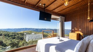 A bedroom with a large bed, wooden ceiling, and a TV. It has a view of mountains and greenery. - Cabana romântica vista incrível VST002 (Anitápolis)