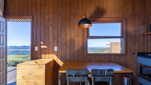 A wooden interior with a dining table and chairs, a hanging pendant light, and large windows offering a view of the countryside. - Cabana romântica vista incrível VST002 (Anitápolis)