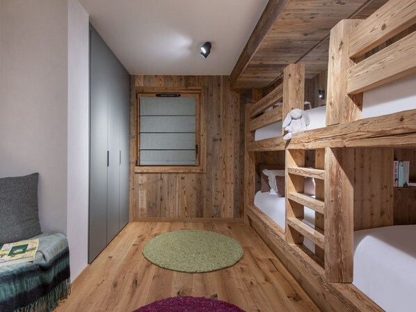 A room with wooden walls, a bunk bed, a window with blinds, a green rug, and a chair with a blanket.