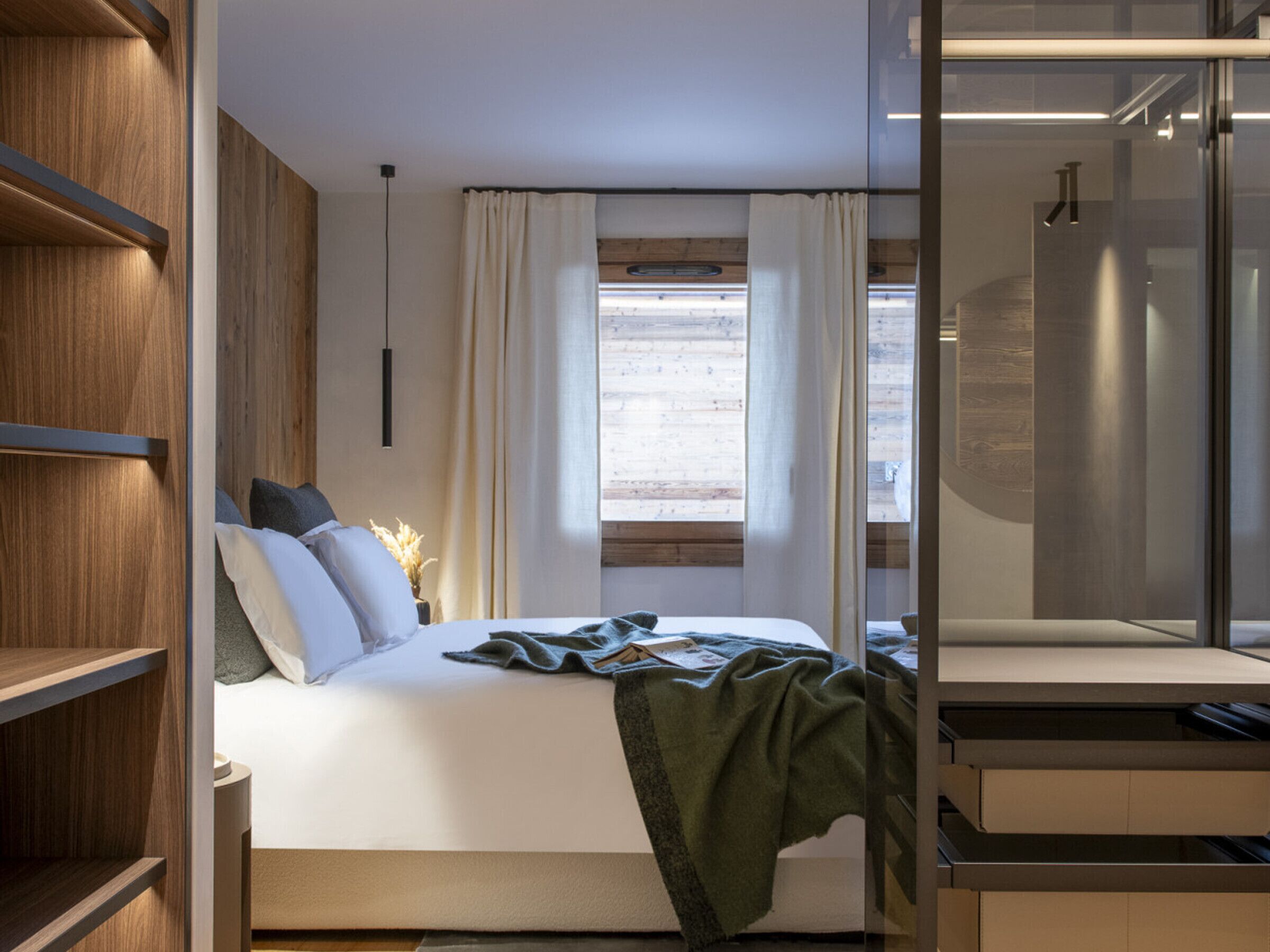 A modern bedroom with a bed, wooden shelving, a window with curtains, and a glass shower area.