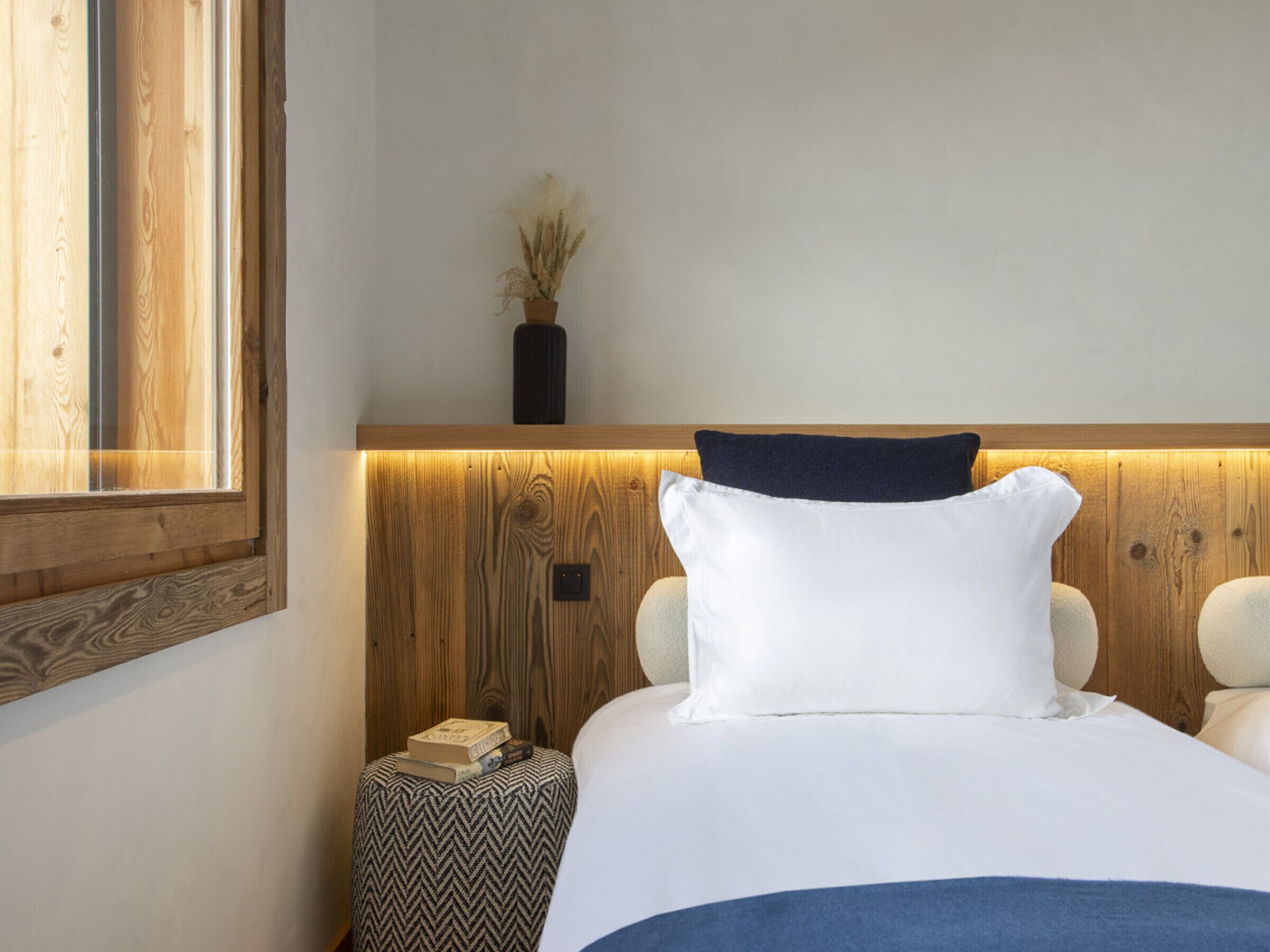 A neatly made bed with white and navy pillows, a wooden headboard, and a small bedside table with books.