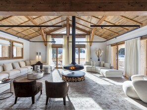 A spacious living room with a high wooden ceiling, a fireplace, and large windows offering a view of the outdoors.
