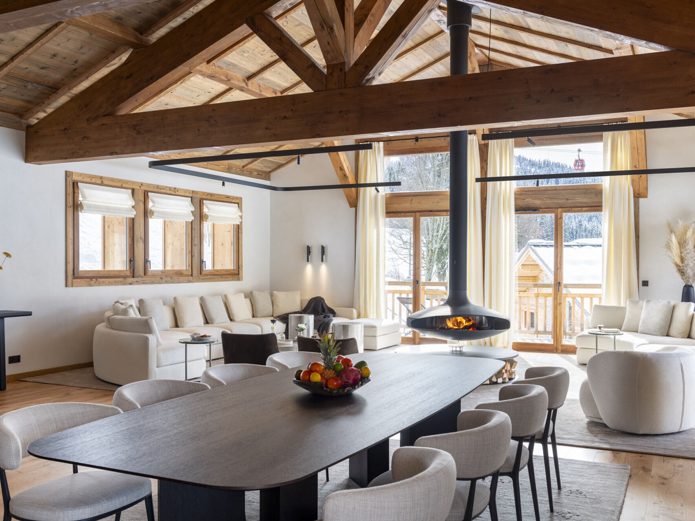 A spacious living area with a large wooden dining table, comfortable seating, and large windows offering a view of the outdoors.