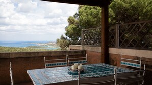 Ein überdachter Außenbereich mit Tisch und Stühlen, mit Blick auf eine reizvolle Landschaft mit Bäumen und Meerblick.