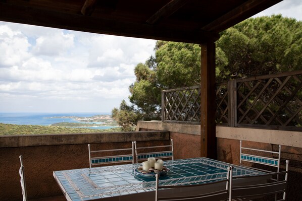 A covered outdoor seating area with a table and chairs, overlooking a scenic landscape with trees and a view of the sea.