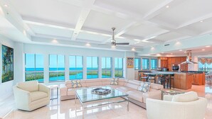 A spacious living room with a large glass coffee table, a sofa set, and a view of the ocean through large windows.