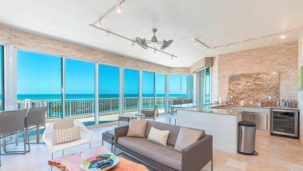 A modern living room with a sofa, chairs, and a coffee table. There is a kitchen area with a sink and a refrigerator. The room has large windows with a view of the ocean.
