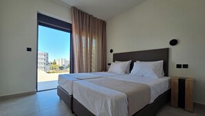 A modern bedroom with a large bed, a view of buildings through a sliding door, and a nightstand with a lamp.
