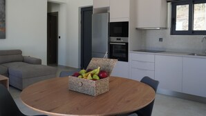 A modern kitchen with a built-in oven, a dining table with a woven fruit basket, and a grey sofa in a living area.