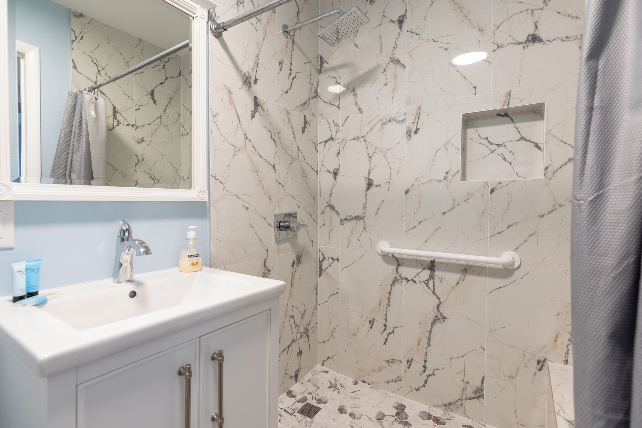 Baño con pared de mármol, lavamanos blanco, espejo, ducha y toallero.
