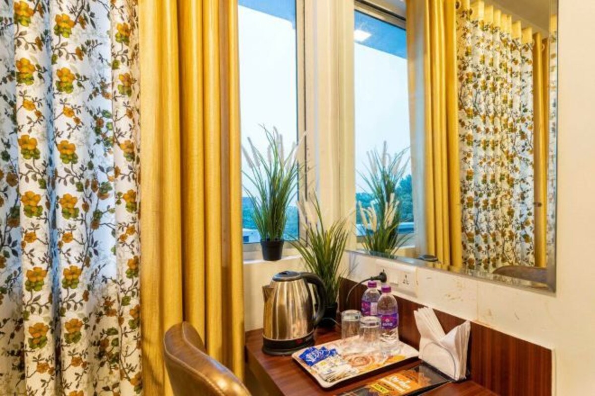 Habitación de hotel con cortinas florales, un escritorio de madera con una bandeja de alimentos para el desayuno, una tetera y una planta en maceta cerca de la ventana.