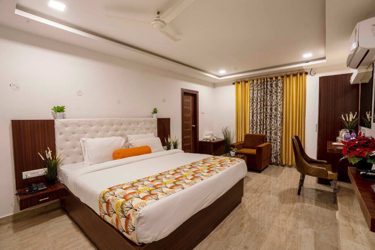 A hotel room with a large bed, a desk with a chair, a small table with a plant, and a ceiling fan.