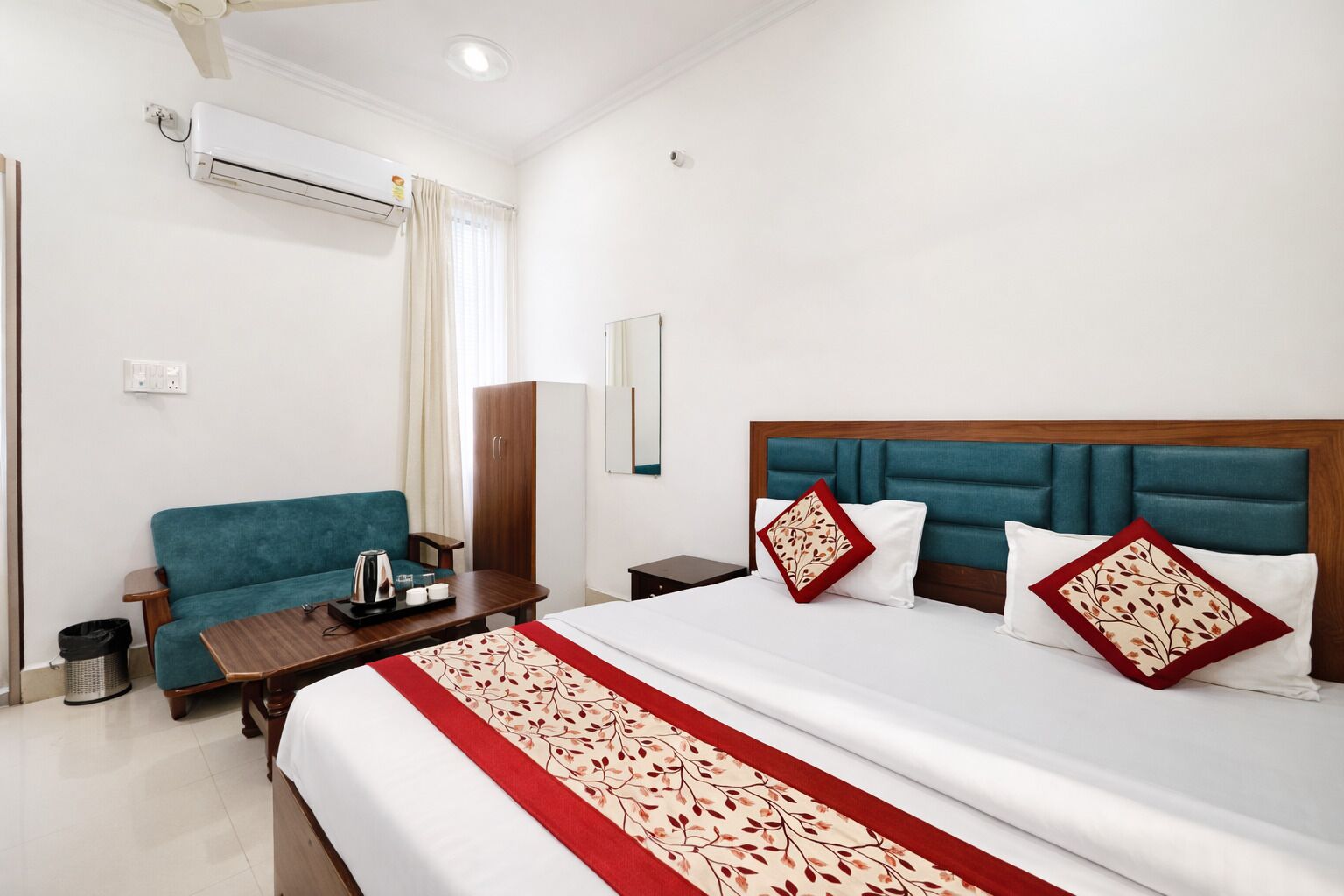 A hotel room with a bed, a sofa, a wooden nightstand, a table with a tray, and a trash bin.