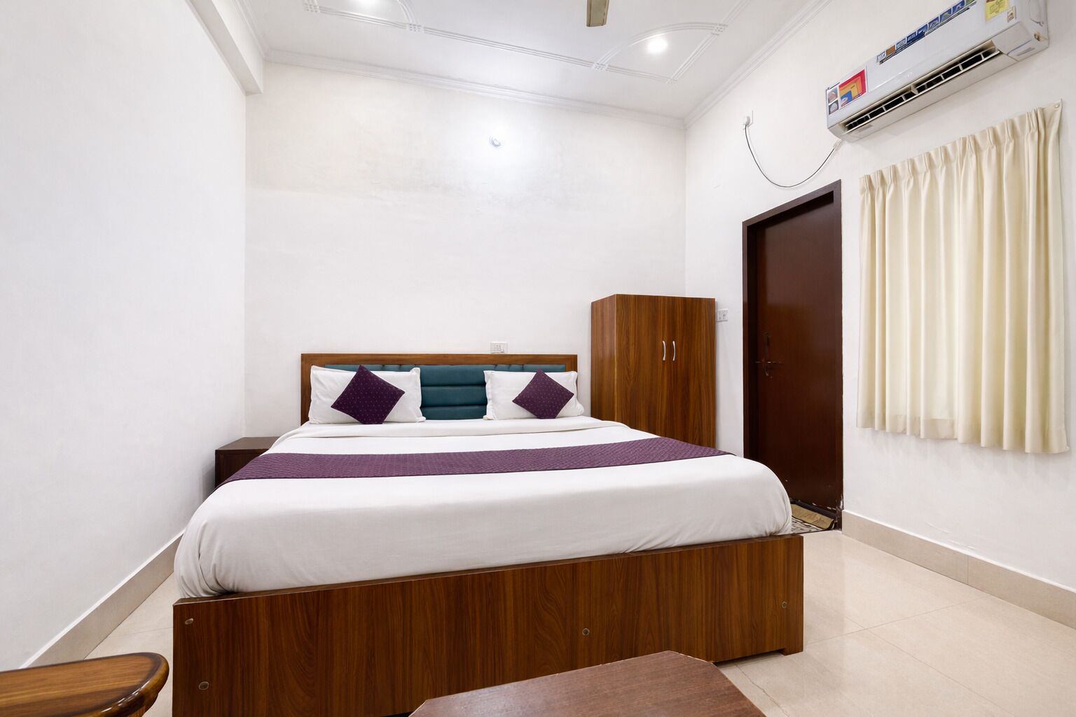 A bedroom with a wooden bed frame, a wardrobe, a bedside table, and a window with curtains.