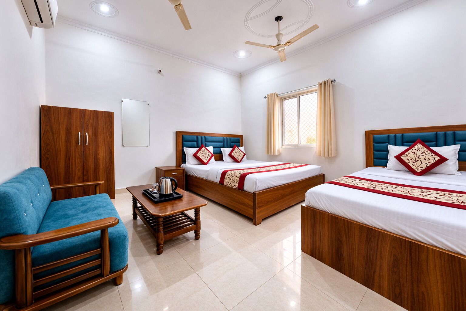 A hotel room with two beds, a wooden wardrobe, a blue sofa, a small wooden table with a kettle, and a ceiling fan.