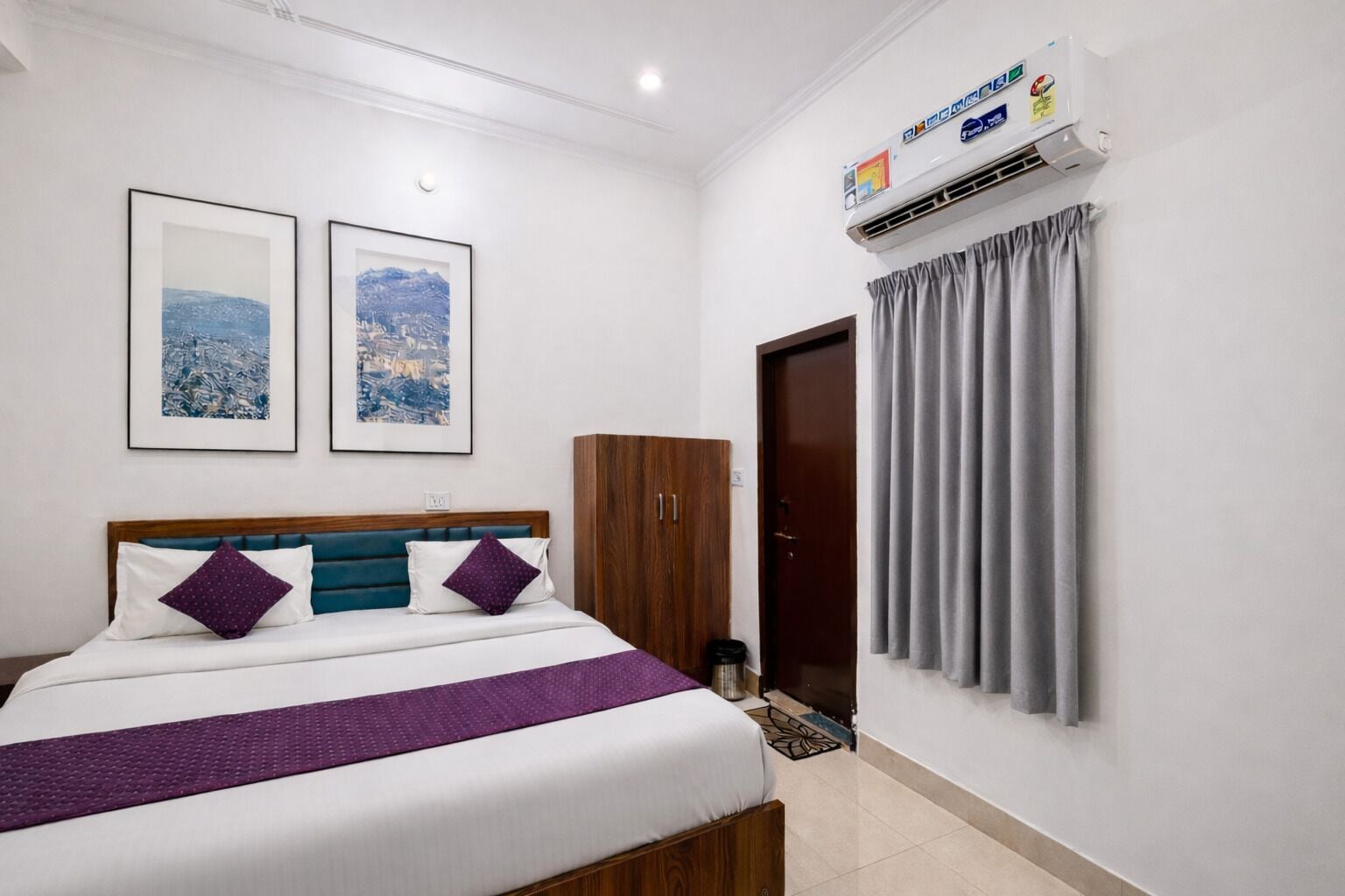 A hotel room with a bed, two framed pictures on the wall, a wardrobe, an air conditioning unit, and a door with curtains.