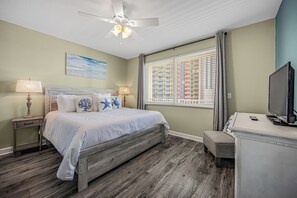 A bedroom with a bed, a ceiling fan, a window with blinds, a television, and a nightstand with a lamp.