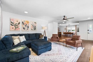 A modern living room with a sectional sofa, a coffee table, and a patterned rug. There are two armchairs and a dining area in the background.