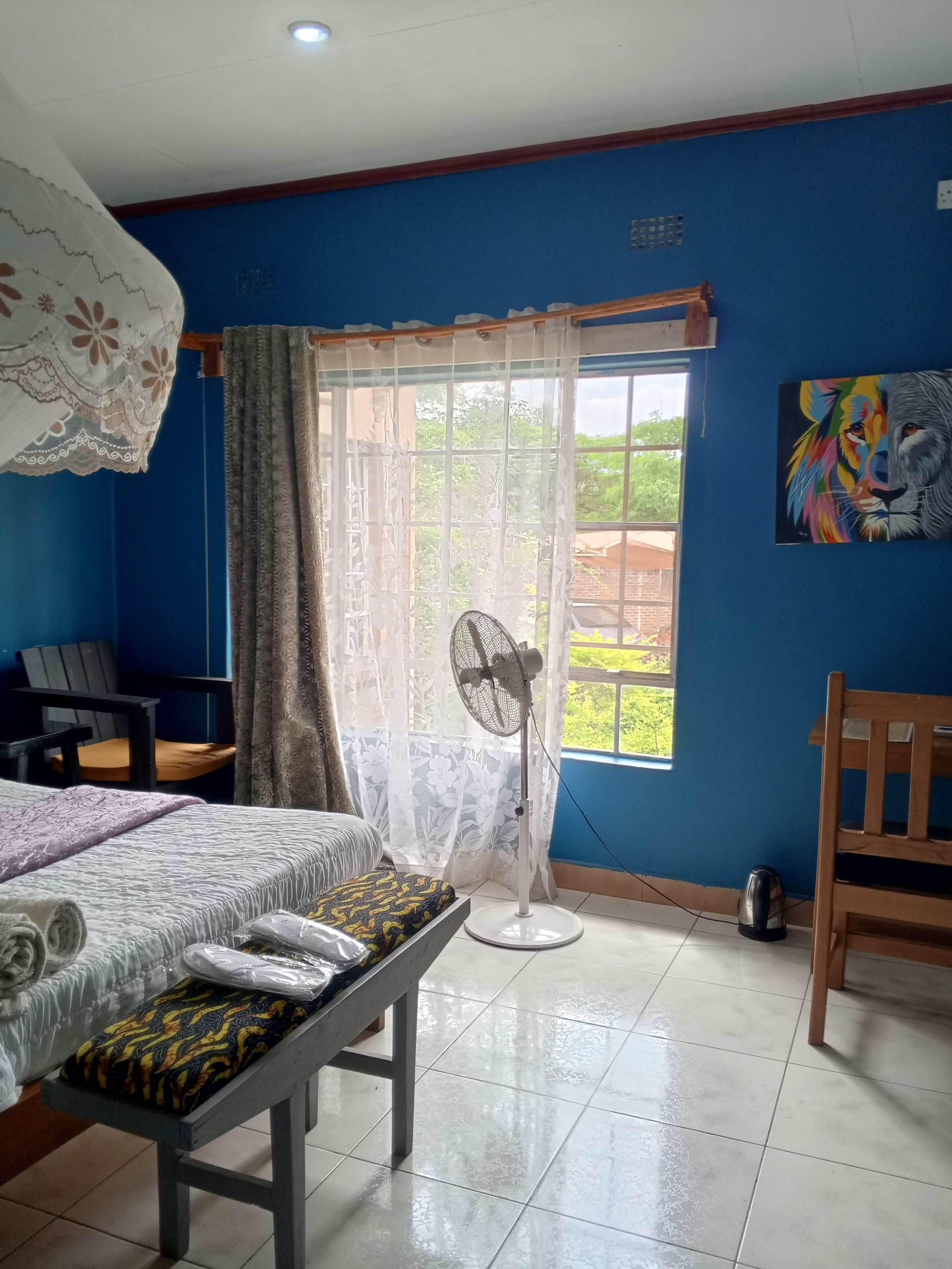 Habitación de hotel con cama, mesa, silla, ventilador, hervidor y ventana con cortinas.