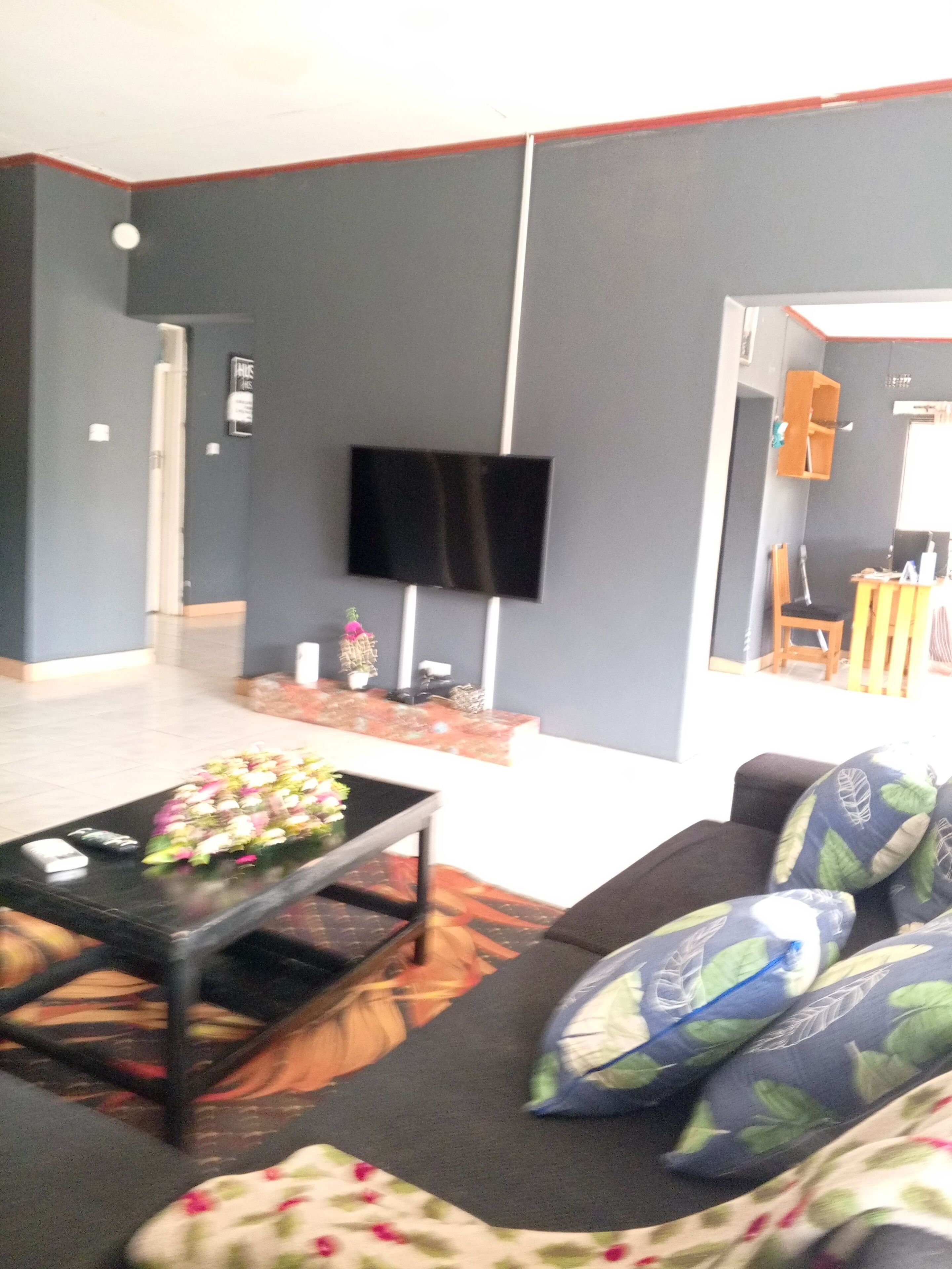 A living room with a flat-screen TV mounted on the wall, a coffee table with a floral arrangement, and a sofa with patterned cushions.