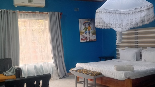 A bedroom with a bed, a desk, a chair, a window with curtains, and a wall-mounted air conditioner. - Quest Lodges and Safaris (Lilongwe)