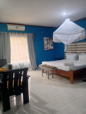 A bedroom with a bed, a desk, a chair, a window with curtains, and a wall-mounted air conditioner. - Quest Lodges and Safaris (Lilongwe)