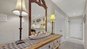 A hallway with a checkered floor, a mirror, two lamps, a wooden counter with a bowl of decorative items, and a door.