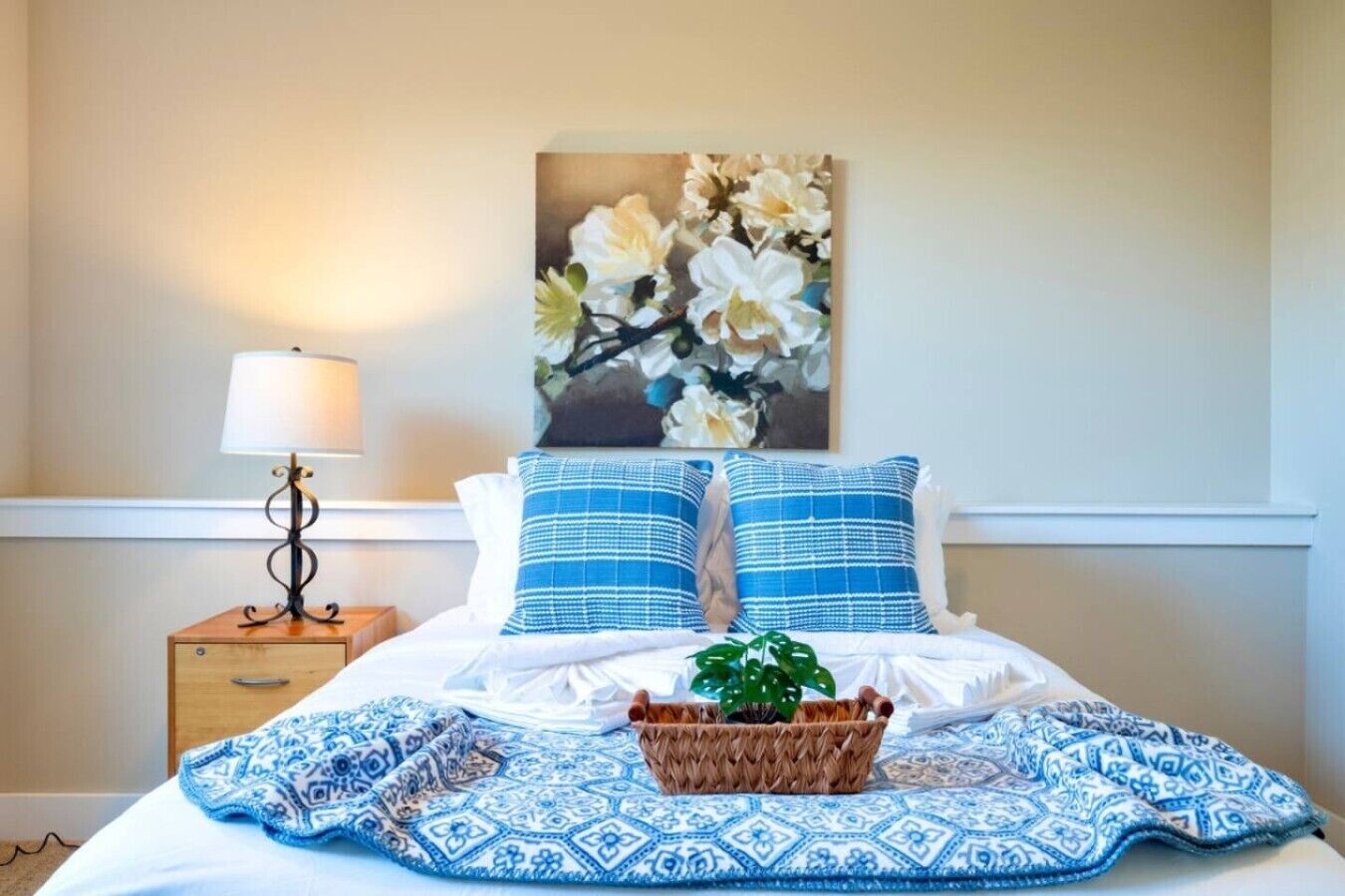 Un letto ben rifatto con biancheria azzurra e bianca, una lampada da comodino, un quadro con dei fiori e un cesto intrecciato con una pianta.