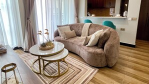 Una sala moderna con un sofá marrón, una mesa de centro redonda y una mesa de comedor con sillas. Cuenta con una zona de cocina con armarios de madera y un horno empotrado. El ambiente tiene suelo de madera y ventanas grandes con cortinas.