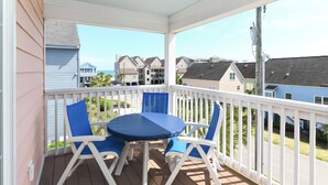 A covered outdoor area with a table and four chairs, a white railing, and a view of houses and the ocean in the background.