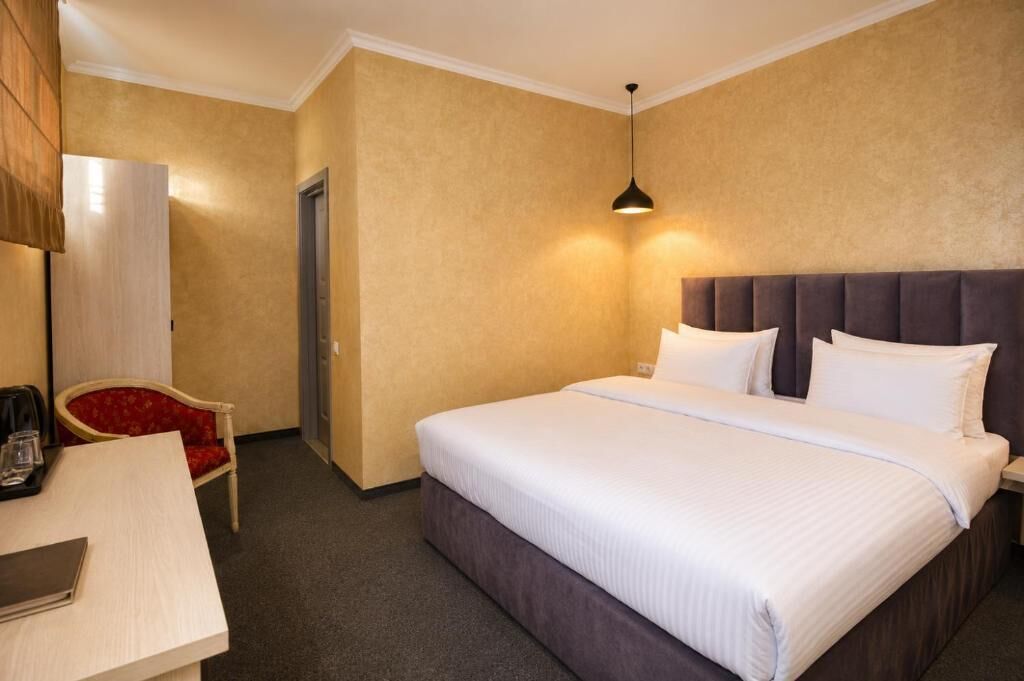 Hotellihuone, jossa on sänky, yöpöytä, tuoli ja riippuvalaisin.