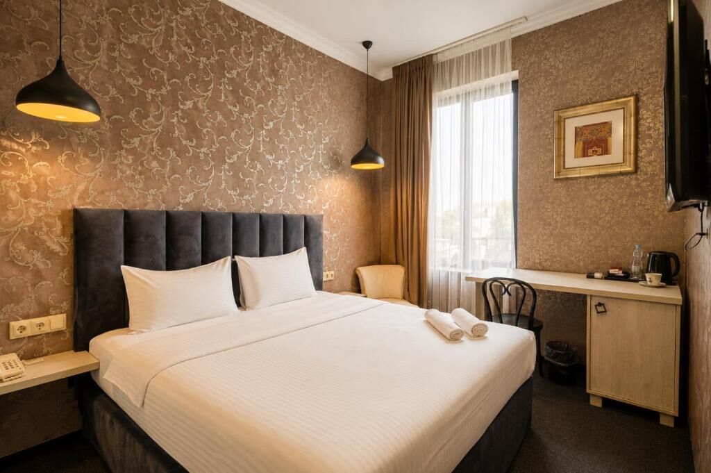 Hotellihuone, jossa on suuri sänky, kaksi riippuvalaisinta, työpöytä tuolilla, televisio ja seinällä oleva kehystetty taulu.